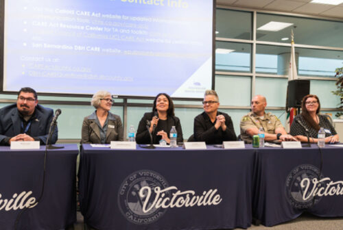 Panelists speak at a CARE Act community information session in Victorville with a presentation screen and audience discussion.