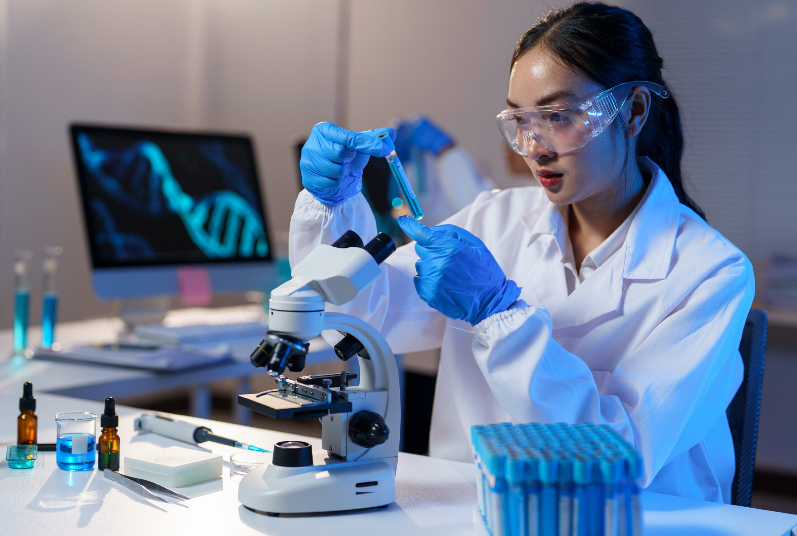 Young female scientist wearing lab coat and gloves carefully examining test tube with blue liquid, conducting research in modern laboratory with microscope and computer displaying